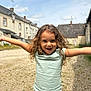 child, smiling, curly_hair, outdoor, sunny, arms_outstretched, happy, street, houses, sky, casual_clothing, young, light_blue_shirt, jeans, playful, daylight, gravel_ground, person, portrait, joy