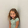 child, smile, curly_hair, sitting, corner, white_wall, shirt, shorts, casual_clothing, portrait, indoor, happy, young, person, cute, playful, light, texture, skin, face