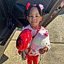 child, girl, balloon, plush_toy, car_door, outdoor, sunlight, red_clothing, heart_pattern, hair_bows, smile, shoes, sidewalk, socks, toy, person, cute, holding, fashion, happy