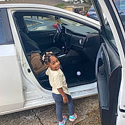 Chanel is registered to the contest to win money with this photo: backpack, car, car_seat, casual_clothing, child, concrete, daylight, hair_bows, happy, open_door, outdoor, parking_lot, person, smile, sneakers, street, toddler, toy_ball, vehicle_interior, window