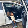 backpack, car, car_seat, casual_clothing, child, concrete, daylight, hair_bows, happy, open_door, outdoor, parking_lot, person, smile, sneakers, street, toddler, toy_ball, vehicle_interior, window