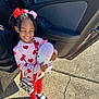 child, girl, smiling, plush_toy, car_door, outdoor, sunlight, red_bows, heart_pattern, sweater, leggings, shoes, white_socks, purse, concrete, happy, cute, standing, accessories, daytime