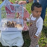 child, toddler, older_child, adult, birdcage, bird, outdoor, grass, garden, family, pet, animal, standing, floral_shirt, overalls, sneakers, sunlight, backyard, portrait, supervision