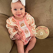 Alexandra is registered to the contest to win money with this photo: baby, infant, smile, girl, headband, bow, pink_clothing, gingerbread, couch, wooden_sign, 5_month, sitting, cute, happy, indoors, child, person, portrait, baby_girl, soft_fabric