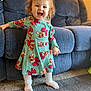 toddler, child, smiling, dress, reindeer_pattern, socks, carpet, couch, indoor, happy, standing, curly_hair, furniture, home, playful, person, cute, cozy, bright, portrait
