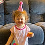 Milani is registered to the contest to win money with this photo: toddler, child, party_hat, pink, smiling, toy, orange, couch, indoor, happy, birthday, dress, balloons_print, furniture, cute, young_child, celebration, person, headwear, living_room