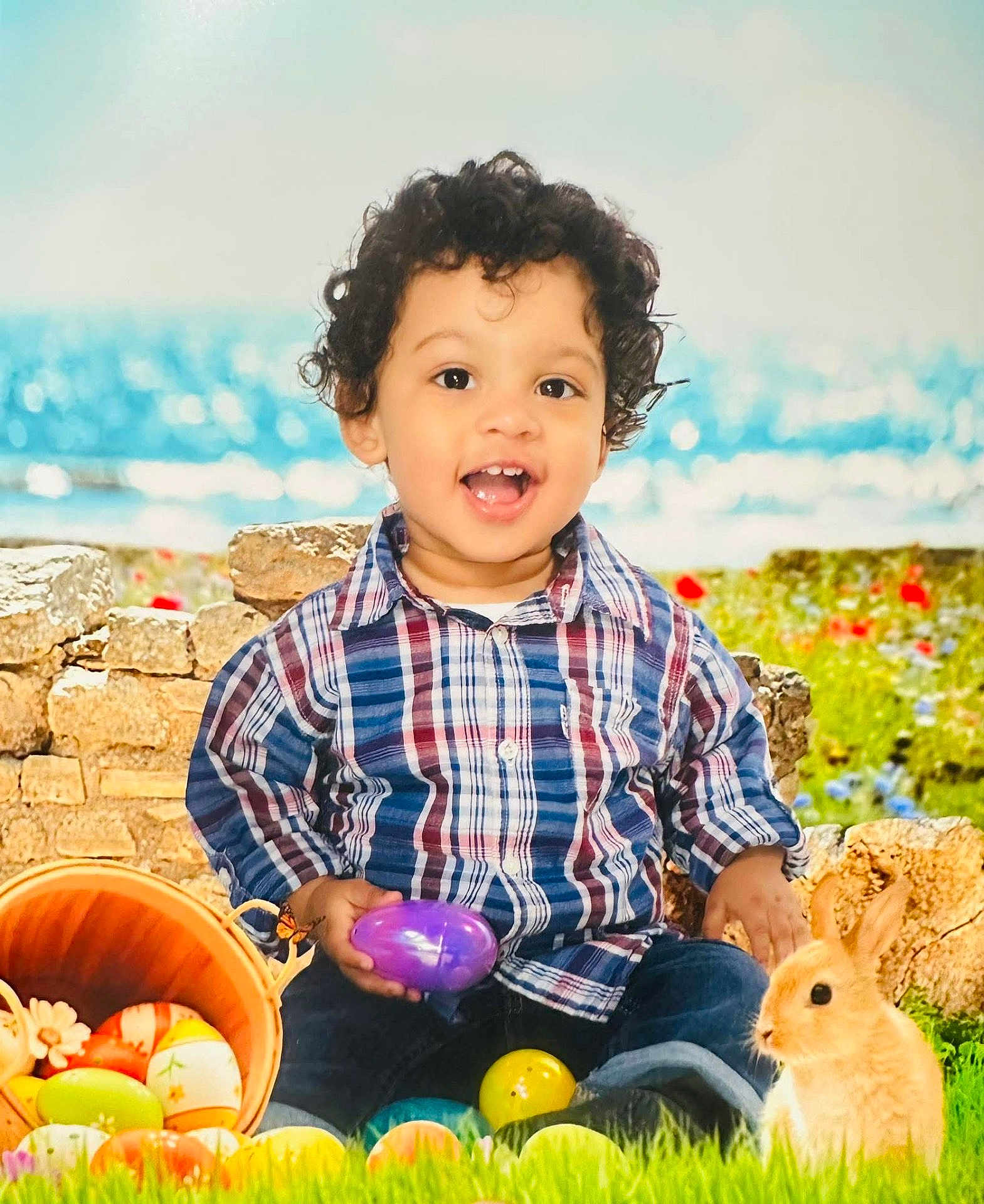 Xavier is registered to the contest to win money with this photo: child, toddler, curly_hair, smiling, easter_eggs, basket, bunny, grass, outdoor, plaid_shirt, blue_jeans, spring, flower, stone_wall, nature, holiday, cute, happy, toy, play