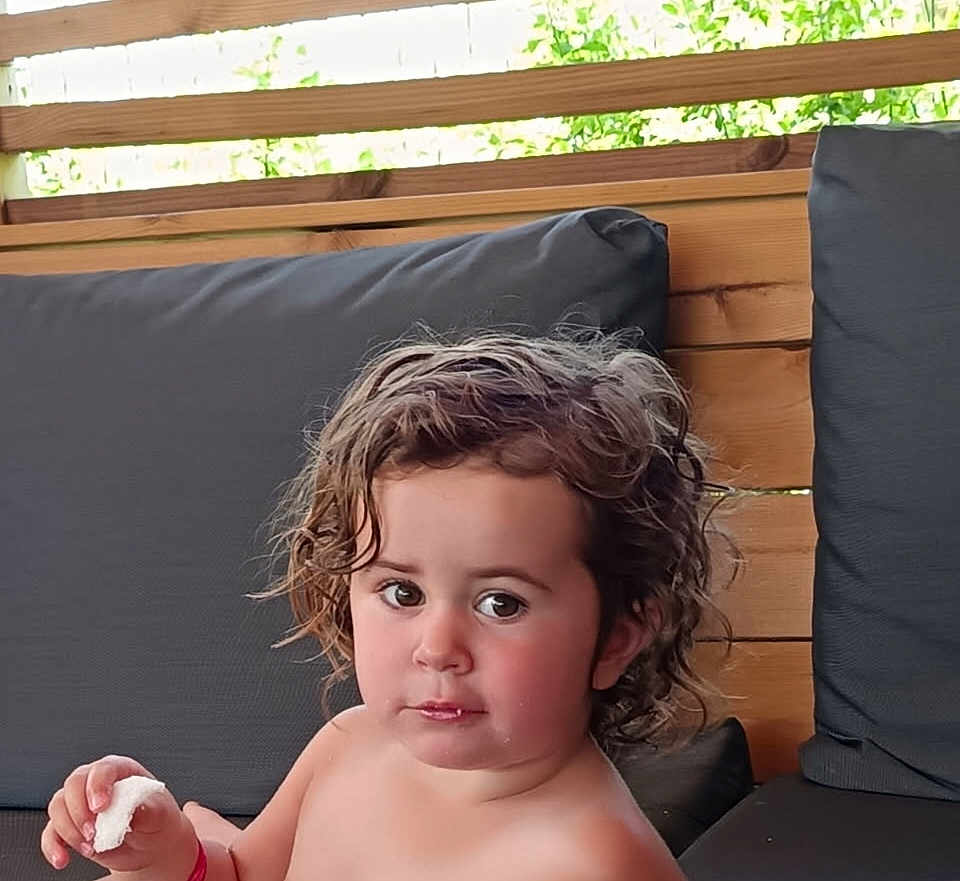 Lola participe au concours pour gagner de l'argent avec cette photo : child, toddler, curly_hair, face, outdoor, bench, cushion, wood, food, snack, expression, person, seat, relaxing, summer, natural_light, casual, young_child, portrait, bare_chest