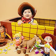 Liam is registered to the contest to win money with this photo: child, baby, toddler, cowboy_hat, costume, box, toys, woody_doll, jessie_doll, buzz_lightyear, mr_potato_head, toy_horse, smile, playful, studio_background, yellow_grid, red_bandana, plush_toy, portrait, happy