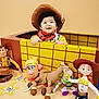 child, baby, toddler, cowboy_hat, costume, box, toys, woody_doll, jessie_doll, buzz_lightyear, mr_potato_head, toy_horse, smile, playful, studio_background, yellow_grid, red_bandana, plush_toy, portrait, happy