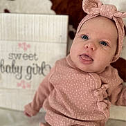 Iris joined the competition — help win amazing prizes! baby, infant, pink_clothing, headband, tongue_out, lying_down, blanket, pattern, sign, text, cute, portrait, child, soft_texture, warm_colors, indoor, happy, face, newborn, smile