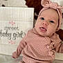 baby, infant, pink_clothing, headband, tongue_out, lying_down, blanket, pattern, sign, text, cute, portrait, child, soft_texture, warm_colors, indoor, happy, face, newborn, smile