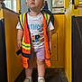 birthday_shirt, child, clothing, curious, expression, floor, indoor, industrial, lighting, person, pipe, play_area, safety_vest, shoes, shorts, socks, standing, toddler, window, yellow