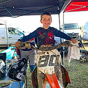Leo a rejoint le concours — aidez-le/la à gagner de superbes lots ! child, boy, smiling, dirt_bike, motorcycle, helmet, canopy, tent, muddy, grass, number_plate, handlebars, racing_gear, van, vehicle, outdoor, happy, portrait, sports, motocross