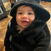 Xander is registered to the contest to win money with this photo: baby, child, hoodie, black_clothing, smiling, happy, indoor, cute, face, eyes, button, furniture, floor, person, portrait, warm_lighting, cozy, infant, young_child, clothing