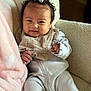 baby, infant, child, onesie, bib, bow, curly_hair, chair, blanket, soft_texture, cozy, indoor, portrait, cute, smile, fingers, clothing, person, warm, relaxed