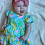 baby, child, infant, headband, bow, dress, floral_pattern, tongue_out, blanket, cute, playful, lying_down, soft_texture, skin, blue_eyes, pink, green, yellow, smile, adorable
