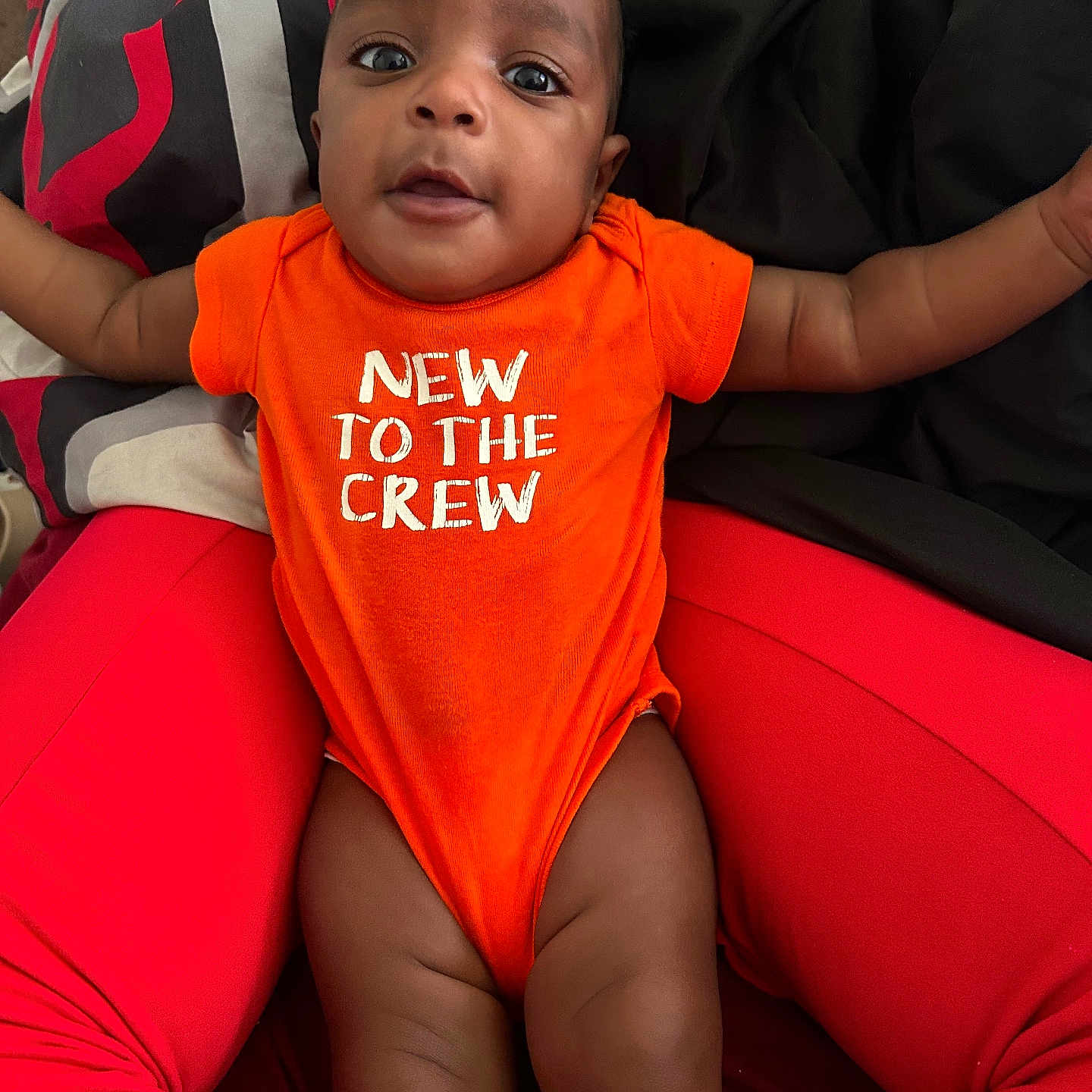 Khalid joined the competition — help win amazing prizes! arms, baby, blanket, casual, child, closeup, comfortable, cute, expression, face, happy, indoor, infant, legs, lying_down, orange_onesie, person, red_pants, skin, text_on_clothing