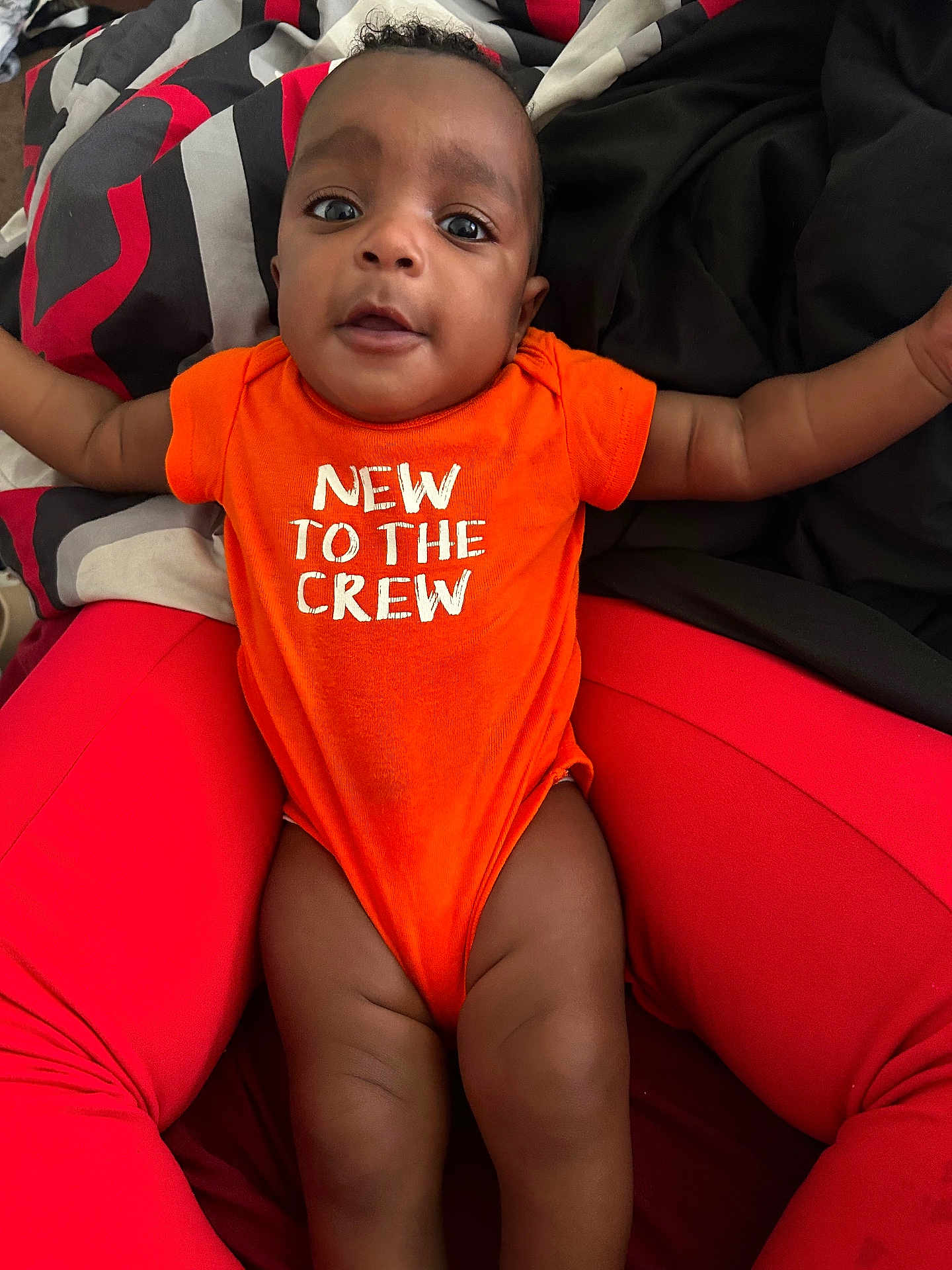Khalid joined the competition — help win amazing prizes! baby, child, infant, orange_onesie, red_pants, person, face, legs, arms, blanket, text_on_clothing, closeup, indoor, comfortable, lying_down, cute, happy, skin, expression, casual