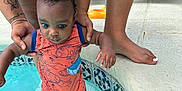 Khalid is registered to the contest to win money with this photo: toddler, child, baby, pool, water, adult, hands, tattoo, orange_clothing, swimwear, outdoor, concrete, tiles, feet, person, sitting, summer, curly_hair, skin, support