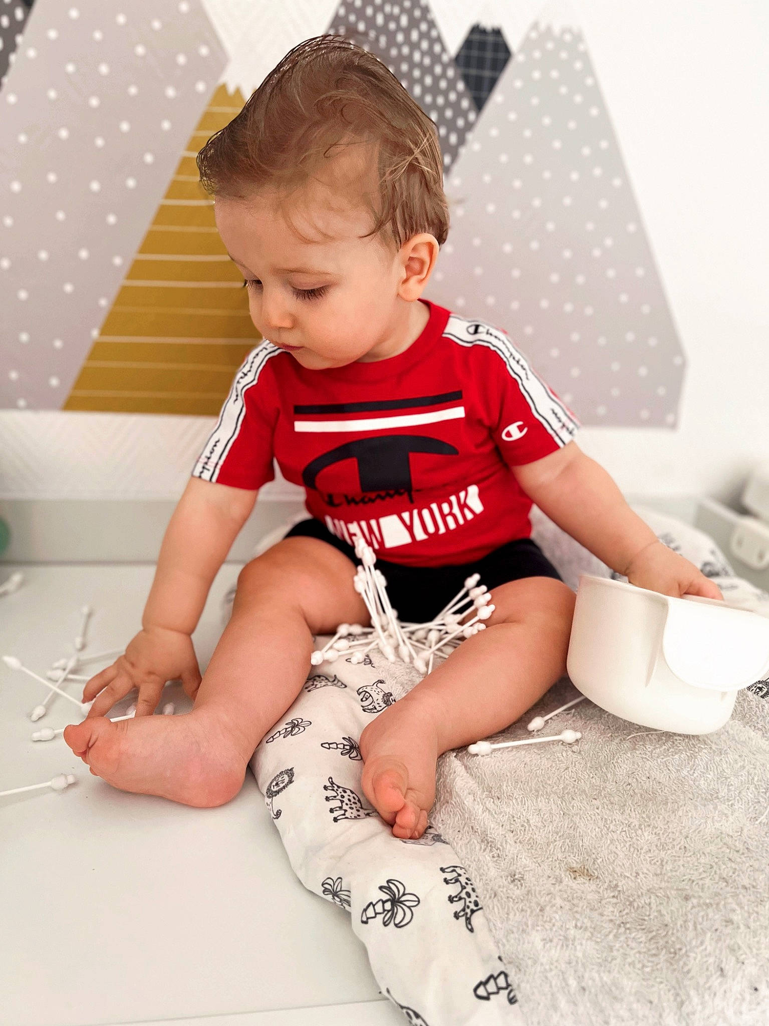 Livio a rejoint le concours — aidez-le/la à gagner de superbes lots ! baby, baby_toddler_clothing, carmine, child, elbow, flooring, foot, fun, human_leg, knee, pattern, person, room, shorts, sitting, sleeve, sock, sportswear, t_shirt, thigh