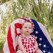 Blair is registered to the contest to win money with this photo: baby, child, infant, portrait, sitting, chair, patriotic, flag, stars, red_white_blue, checkered_dress, headband, outdoor, garden, willow_tree, foliage, backyard, natural_light, smiling, photography