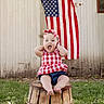 Blair is registered to the contest to win money with this photo: baby, child, infant, portrait, sitting, barrel, wooden_barrel, american_flag, patriotic, outdoor, grass, barn, wooden_wall, red_white_blue, checkered_dress, headband, smiling, surprised, barefoot, country_style