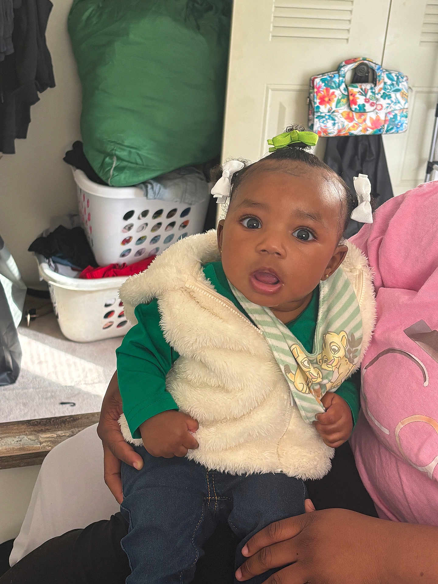 Janiyah is registered to the contest to win money with this photo: baby, child, person, indoor, clothing, vest, jeans, hair_bow, green_shirt, laundry_basket, bag, hand, curious, cute, portrait, seated, expression, casual, young, holding
