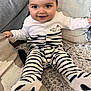 baby, infant, smiling, striped_pajamas, onesie, sitting, couch, cushion, blanket, plush_rug, closeup, portrait, face, big_eyes, chubby_cheeks, hands, feet, socks, home_interior, cozy