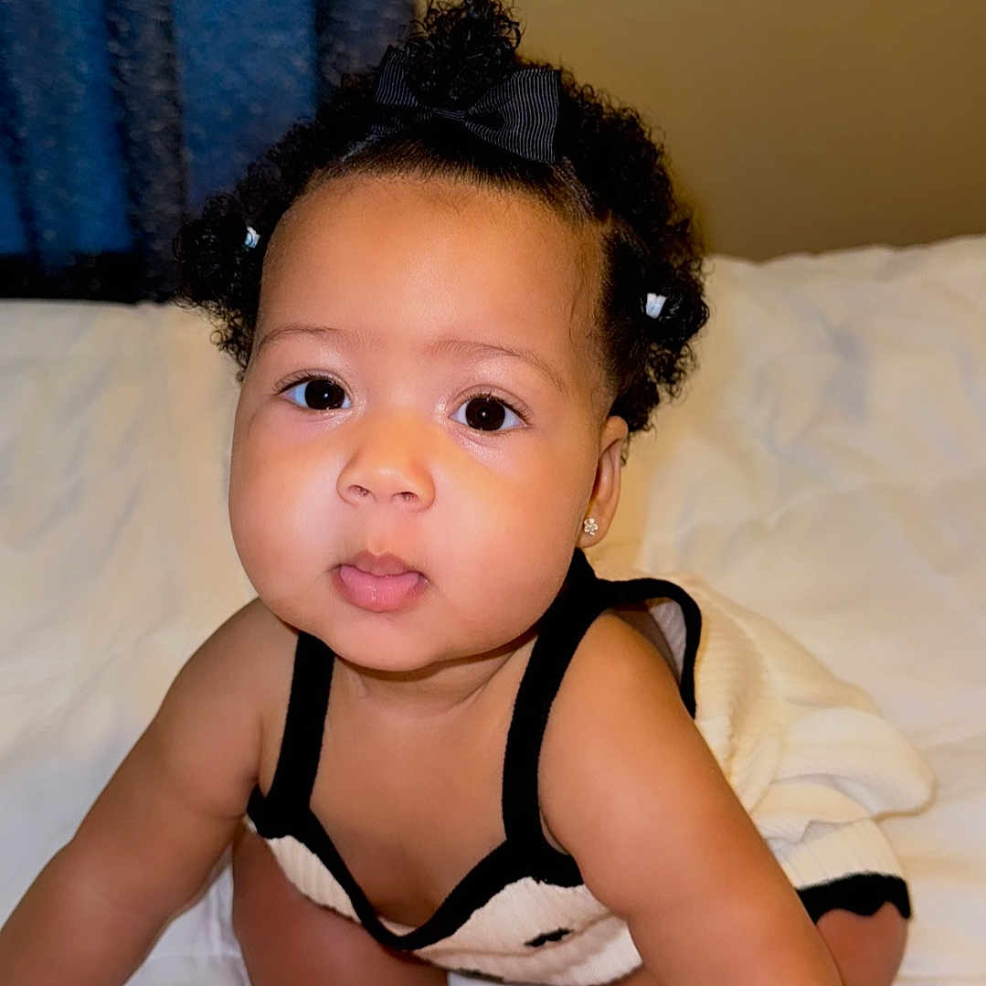 Ramonie is registered to the contest to win money with this photo: baby, bed, black_and_white_clothing, blanket, bow, child, chubby_cheeks, curly_hair, cute, earrings, eyes, face, feet, indoors, infant, person, portrait, sitting, skin, young