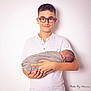 boy, baby, child, newborn, glasses, white_shirt, swaddled, holding, portrait, studio, smile, person, hands, infant, family, sibling, cute, young, indoor, soft_lighting