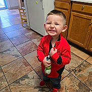 Romeo is registered to the contest to win money with this photo: child, smiling, red_jacket, blue_jeans, red_shoes, kitchen, tile_floor, refrigerator, wooden_cabinets, toy, bottle, happy, indoor, person, young_child, short_hair, grinning, standing, clothing, home