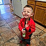 child, smiling, red_jacket, blue_jeans, red_shoes, kitchen, tile_floor, refrigerator, wooden_cabinets, toy, bottle, happy, indoor, person, young_child, short_hair, grinning, standing, clothing, home