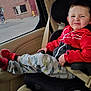 child, car_seat, red_jacket, pants, airplane_print, sneakers, seatbelt, car_interior, window, rain_drops, face, expression, funny_face, sitting, casual, indoor, person, clothing, transportation, daylight
