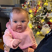 Chloe is registered to the contest to win money with this photo: baby, bib, blue_eyes, cheerful, christmas_tree, closeup, cute, face, festive, hand, holding, holiday_decorations, indoor, infant, lights, ornaments, person, pink_clothing, smiling, warm