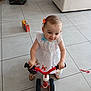 toddler, girl, tricycle, indoor, tile_floor, dress, bow, toy_doll, child, footwear, bag, colorful, smiling, play, baby, floor, hand, furniture, home, person