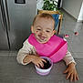 toddler, child, pink_bib, blackberries, food, snack, kitchen, table, face_stains, happy, indoor, plant, chair, refrigerator, tile_floor, footwear, hand, person, young_child, casual_clothing