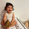 baby, bed, child, curly_hair, cushion, cute, face, furniture, happy, indoor, name_plaque, person, play, plush_toy, sitting, smiling, star_pattern, toddler, wall, white_tank_top
