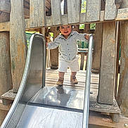Ethan participe au concours pour gagner de l'argent avec cette photo : casual_clothing, child, curly_hair, daylight, fence, fun, hands, metal_slide, nature, outdoor, park, person, play, playground, shoes, slide, smiling, toddler, wood, wooden_structure