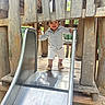 casual_clothing, child, curly_hair, daylight, fence, fun, hands, metal_slide, nature, outdoor, park, person, play, playground, shoes, slide, smiling, toddler, wood, wooden_structure