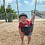 child, toddler, swing, playground, red_shirt, cap, old_navy, smiling, barefoot, wood_chips, outdoor, daylight, sky, clouds, water, pier, railing, greenery, happy, fun
