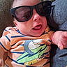 baby, child, sunglasses, striped_shirt, orange_shirt, shark_graphic, couch, smiling, happy, indoor, person, infant, cute, reaching_hand, clothing, portrait, relaxed, head, face, casual