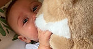 Elijah is registered to the contest to win money with this photo: baby, infant, child, plush_toy, stuffed_animal, dog_toy, face, eyes, smiling, curious, sitting, lying_down, hand, arm, white_clothing, blanket, crib, nursery, indoor, portrait