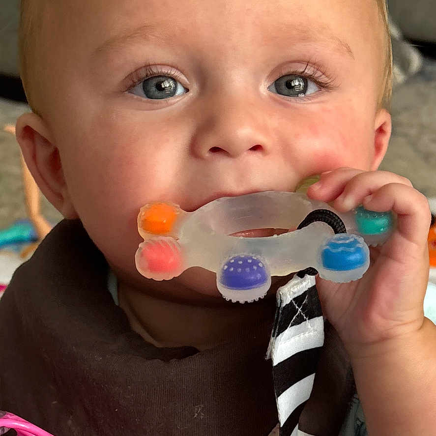 Corbin joined the competition — help win amazing prizes! animal_print, baby, bib, blue_eyes, child, closeup, clothing, comfort, couch, cute, face, hand, indoor, infant, person, pink_toy, play, portrait, striped_ribbon, teething_toy