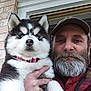 man, dog, puppy, husky, beard, cap, hand, window, brick_wall, outdoor, pet, animal, portrait, fur, blue_eyes, holding, casual_clothing, male, looking_at_camera, closeup