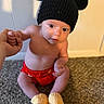 Jordan Jr is registered to the contest to win money with this photo: baby, child, infant, knit_hat, black_hat, red_shorts, yellow_booties, carpet, hand_holding, sitting, indoor, wall, shadow, cute, young_child, portrait, looking_up, bare_chest, human, floor