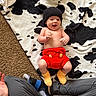 baby, infant, smiling, hat, crocheted_clothing, red_shorts, yellow_booties, black_and_white_blanket, carpet, person, legs, socks, tattoo, hand, happy, lying_down, indoor, cozy, cute, playful