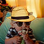 baby, child, stuffed_toy, green_toy, plush, hat, sunglasses, mustache, striped_shirt, indoor, curtains, cushion, blanket, soft_lighting, cute, portrait, sitting, hands, face, playful