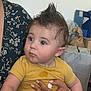 baby, child, person, face, hair, mohawk, yellow_shirt, stain, hand, adult, floral_clothing, indoor, storage, box, curious, portrait, skin, eyes, expression, holding