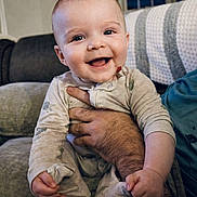 Fionn joined the competition — help win amazing prizes! baby, smiling, infant, couch, hand, adult_arm, onesie, happy, indoor, face, child, cozy, blanket, living_room, portrait, cute, skin, sitting, expression, comfort